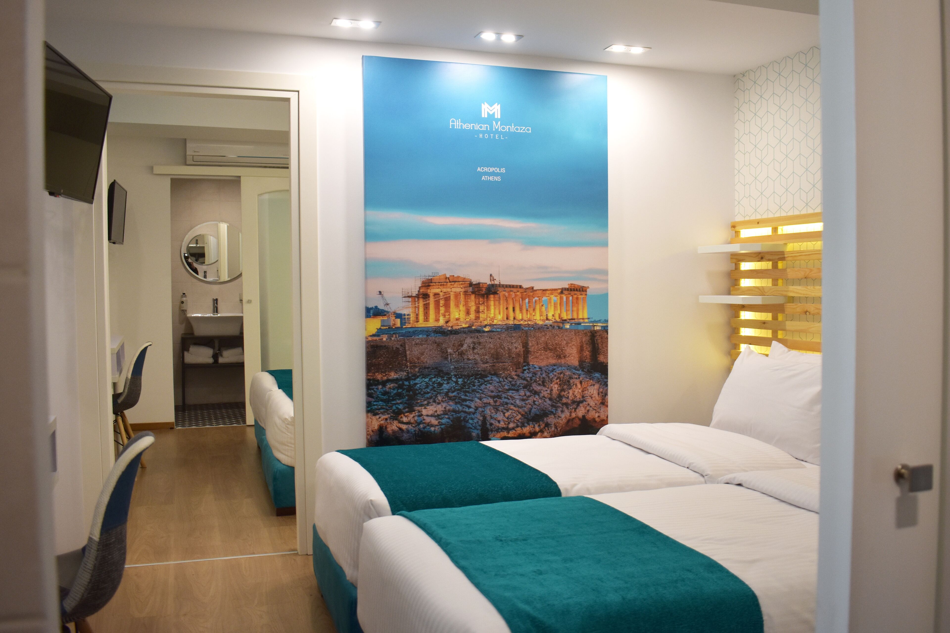 Une chambre d’hôtel avec une grande affiche représentant l’Acropole, un lit avec une literie blanche et turquoise, une tête de lit en bois et un bureau avec une chaise.
