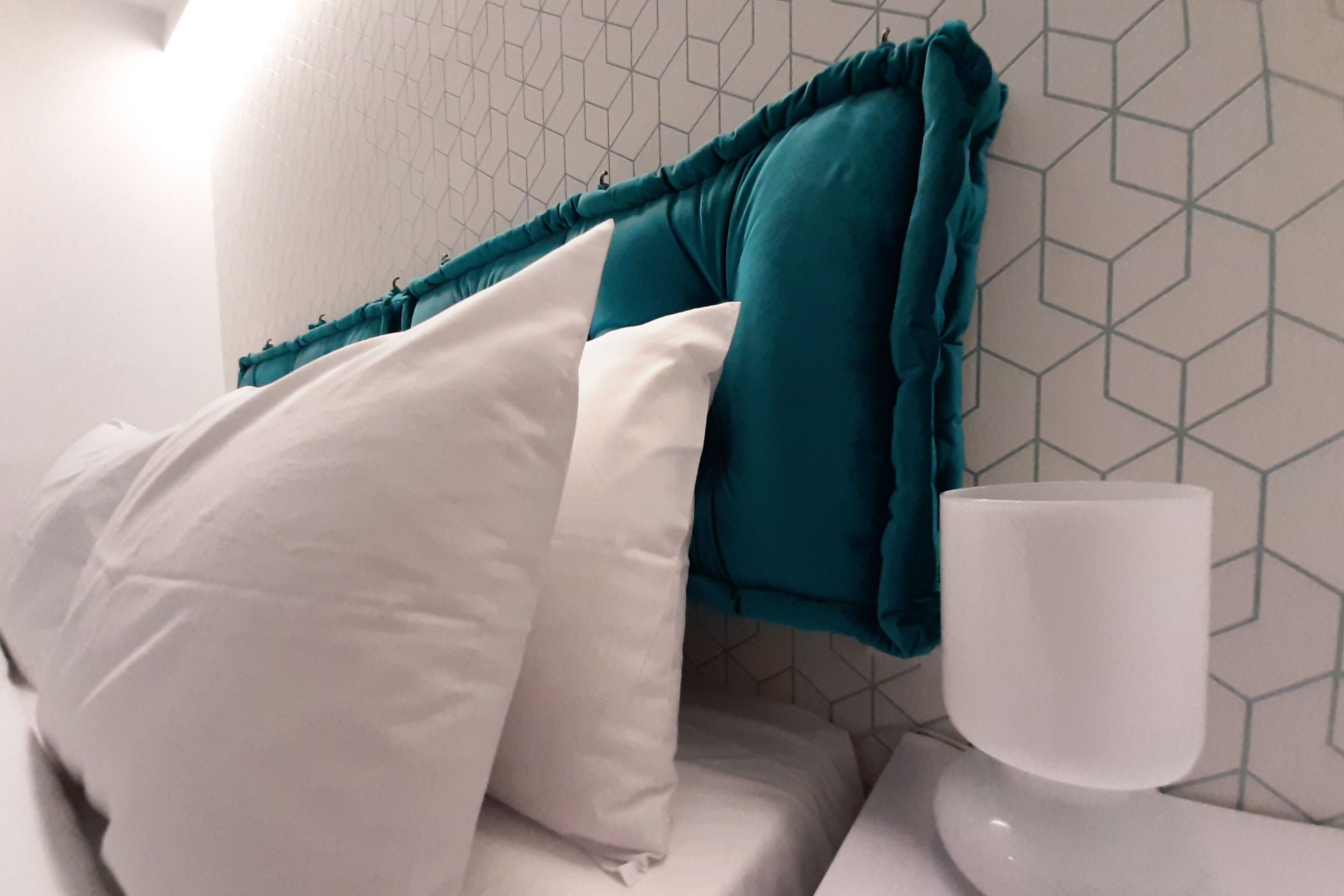 A neatly made bed with white and teal pillows, a white bedside table, and a white cup.
