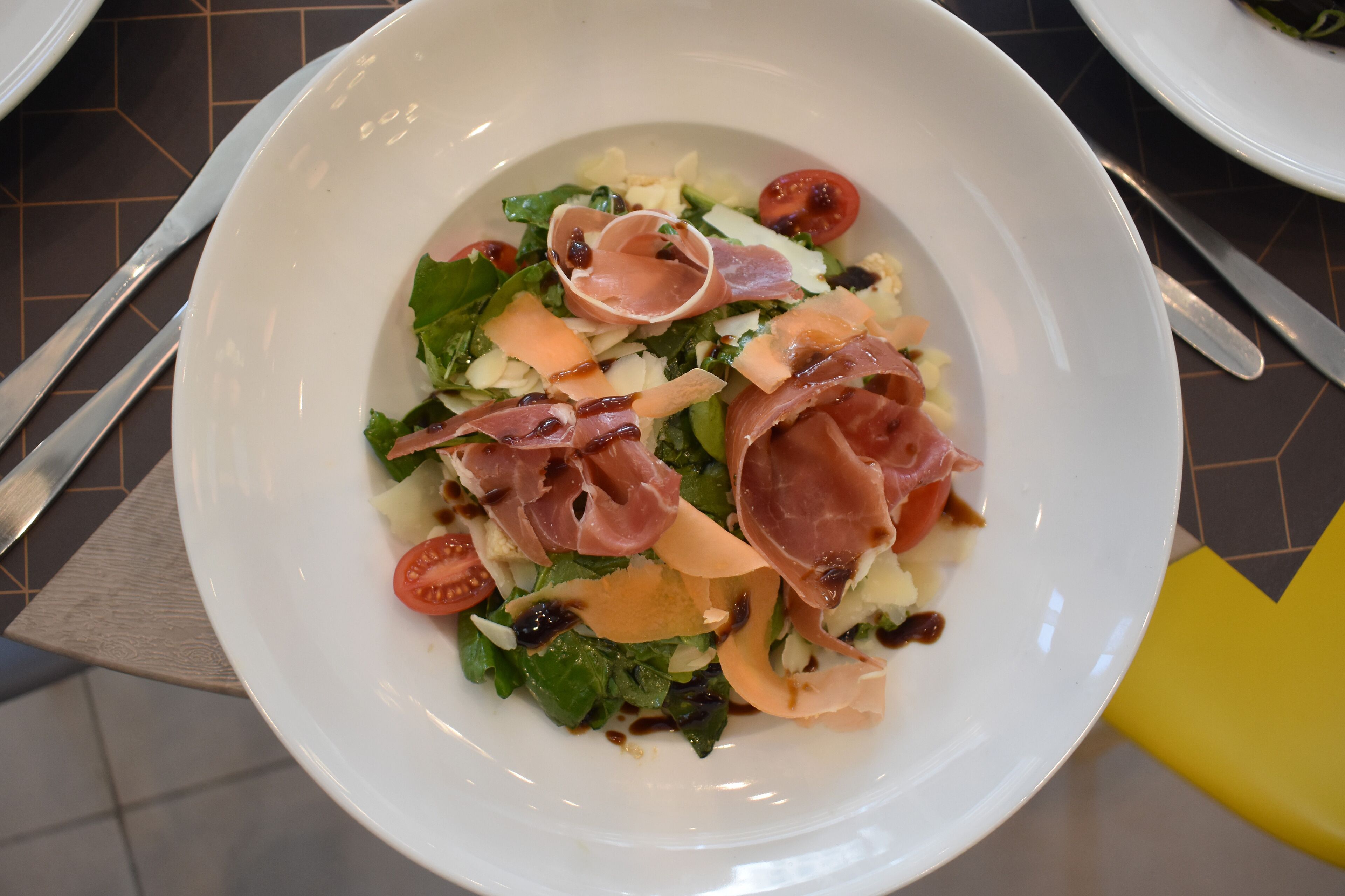 Een witte kom met een salade, groenten, tomaten en parmaham.