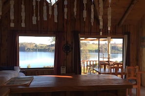 A wooden cabin with a large window overlooking a lake, a wooden table, and a view of the landscape.