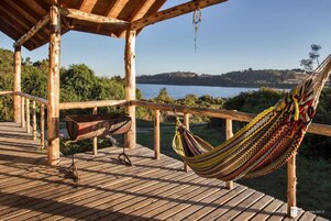 Una terraza de madera con una hamaca y una parrilla, con vistas a un lago y un bosque.