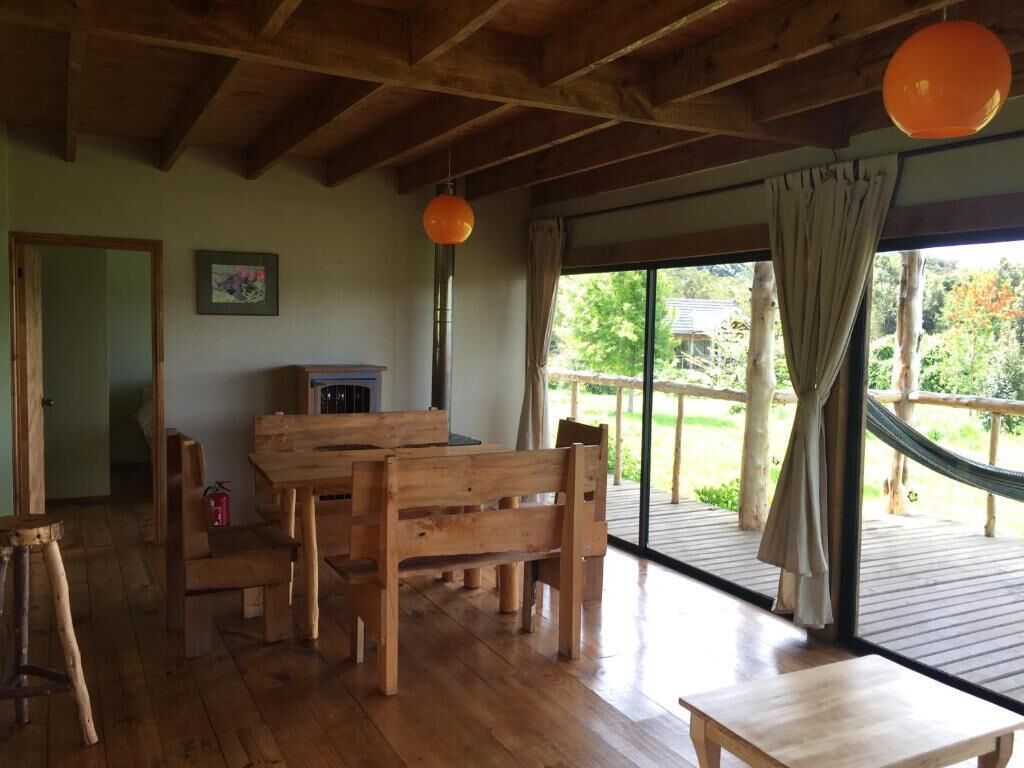 Un coin repas en bois avec une table et des bancs, un luminaire orange suspendu et une vue sur un jardin par le biais d’une grande fenêtre.