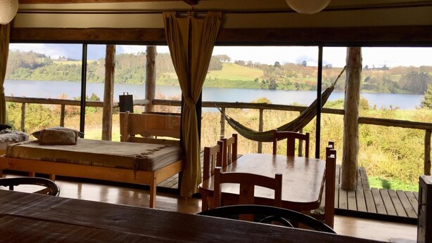 A rustic cabin with a wooden deck, a hammock, a bed, and a table. - Antuquelen Lodge Cabañas (Frutillar)