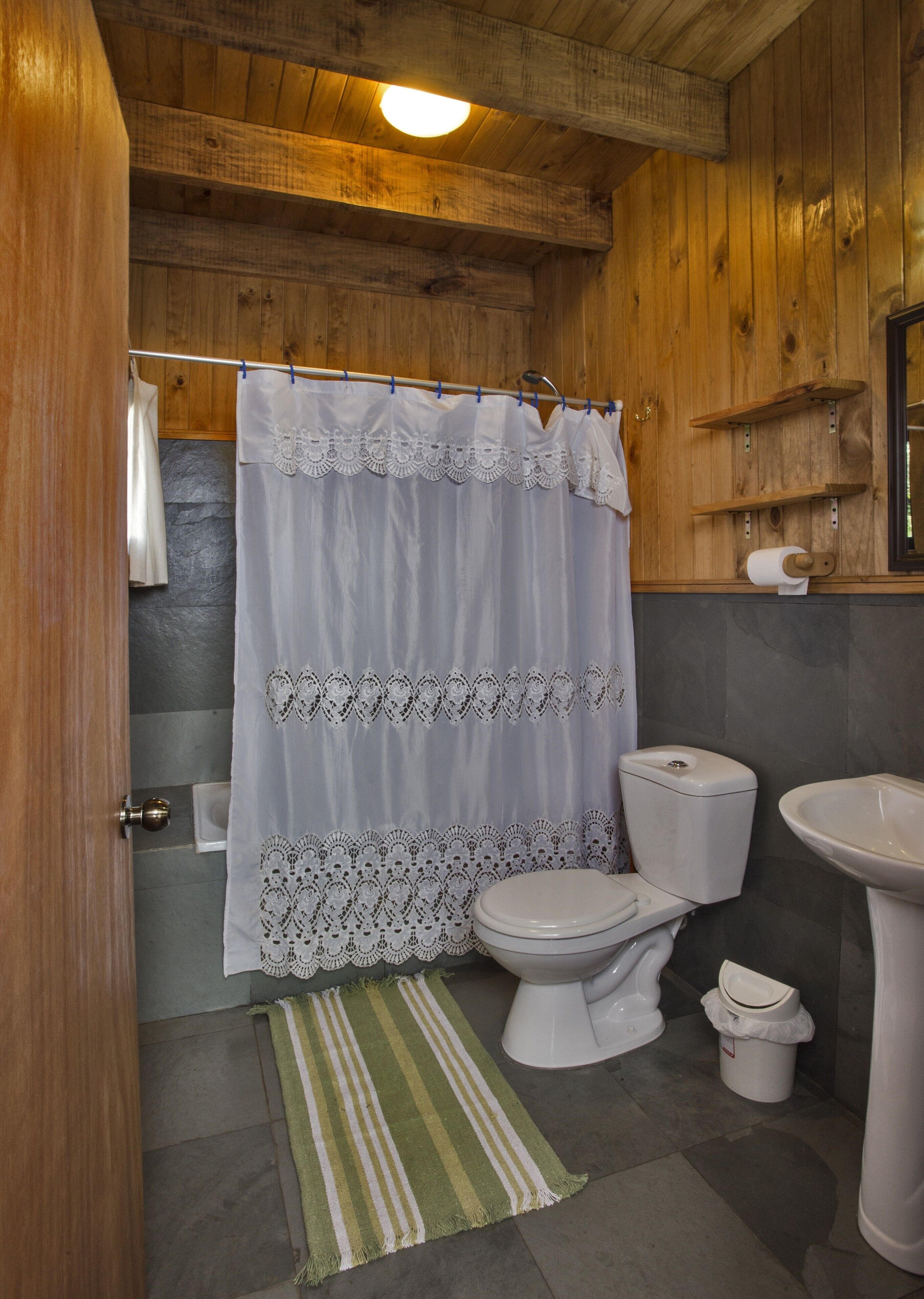 A bathroom with a wooden ceiling, a white toilet, a sink, and a shower curtain with lace detailing.