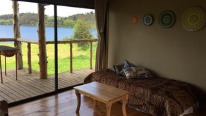 A room with a wooden floor, a sofa with patterned cushions, a wooden table, and a view of a lake through large windows. - Antuquelen Lodge Cabañas (Frutillar)