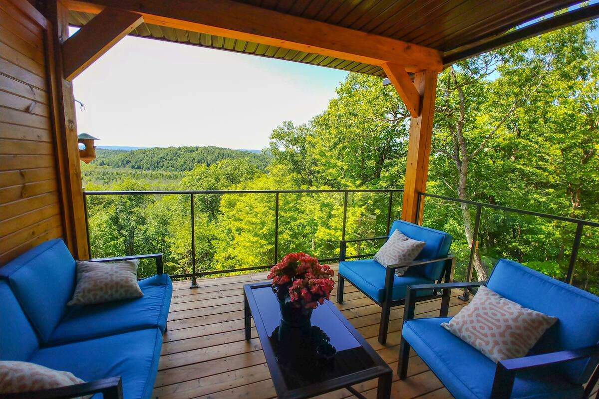 A wooden deck with blue sofas and a table with flowers, overlooking a forested landscape.