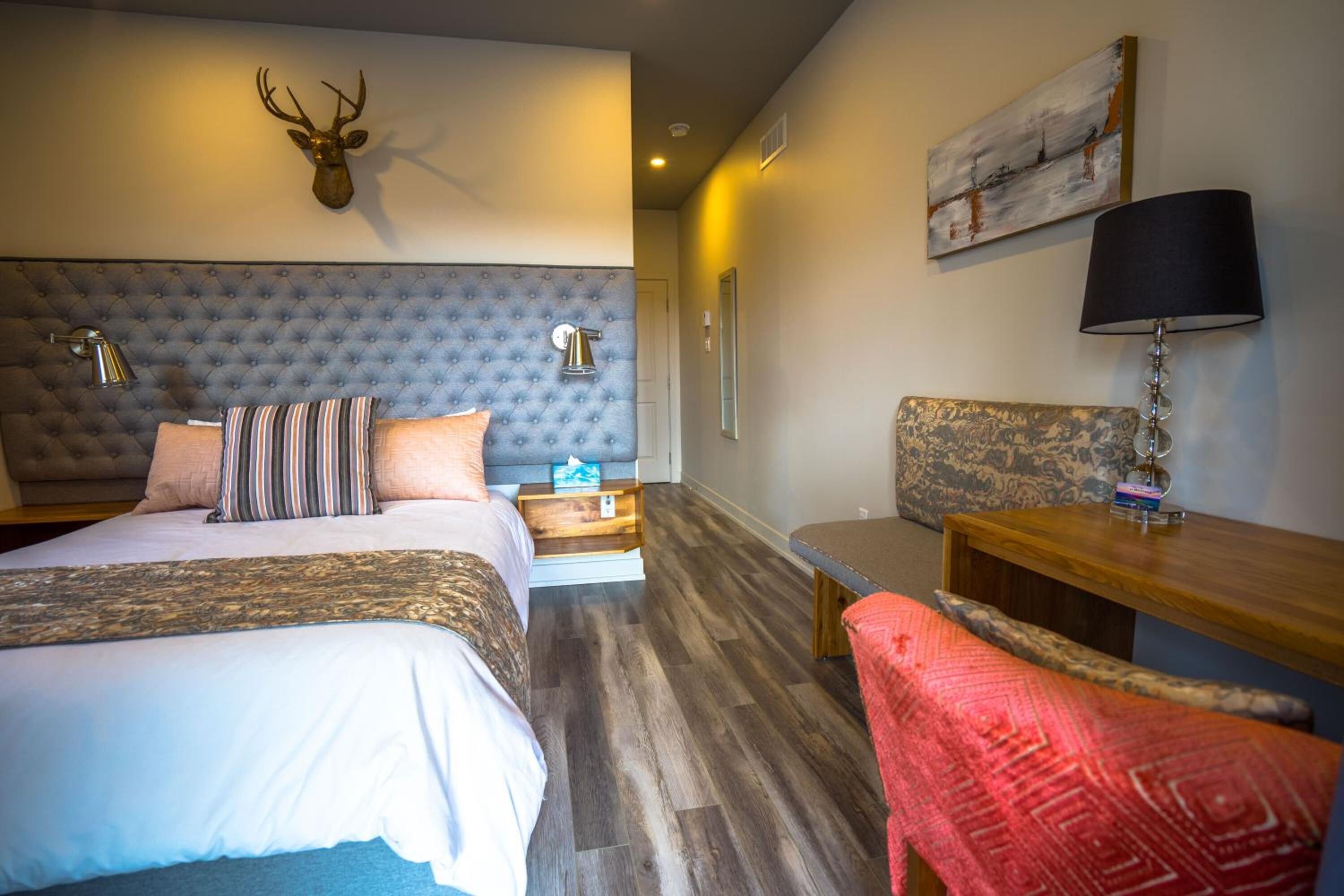 Una habitación de hotel con un cabecero capitonado, una cama con ropa de cama a rayas, una mesita de noche de madera, un escritorio con una lámpara y una manta roja.