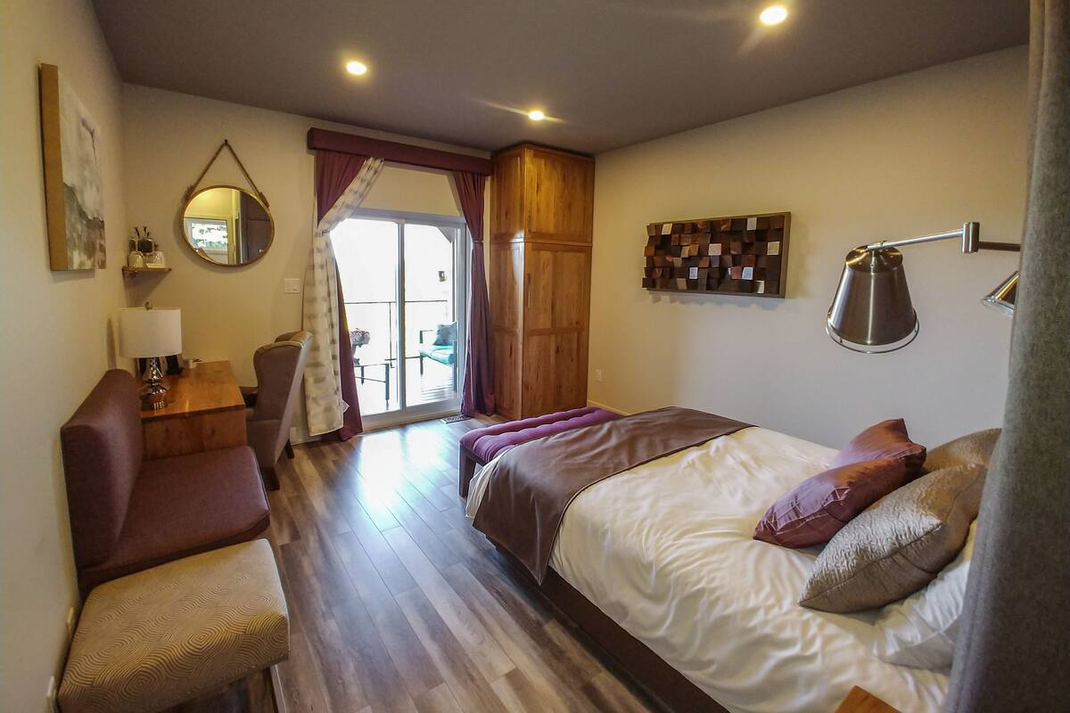 A bedroom with a bed, a desk, a chair, a lamp, a mirror, and a view through a door.