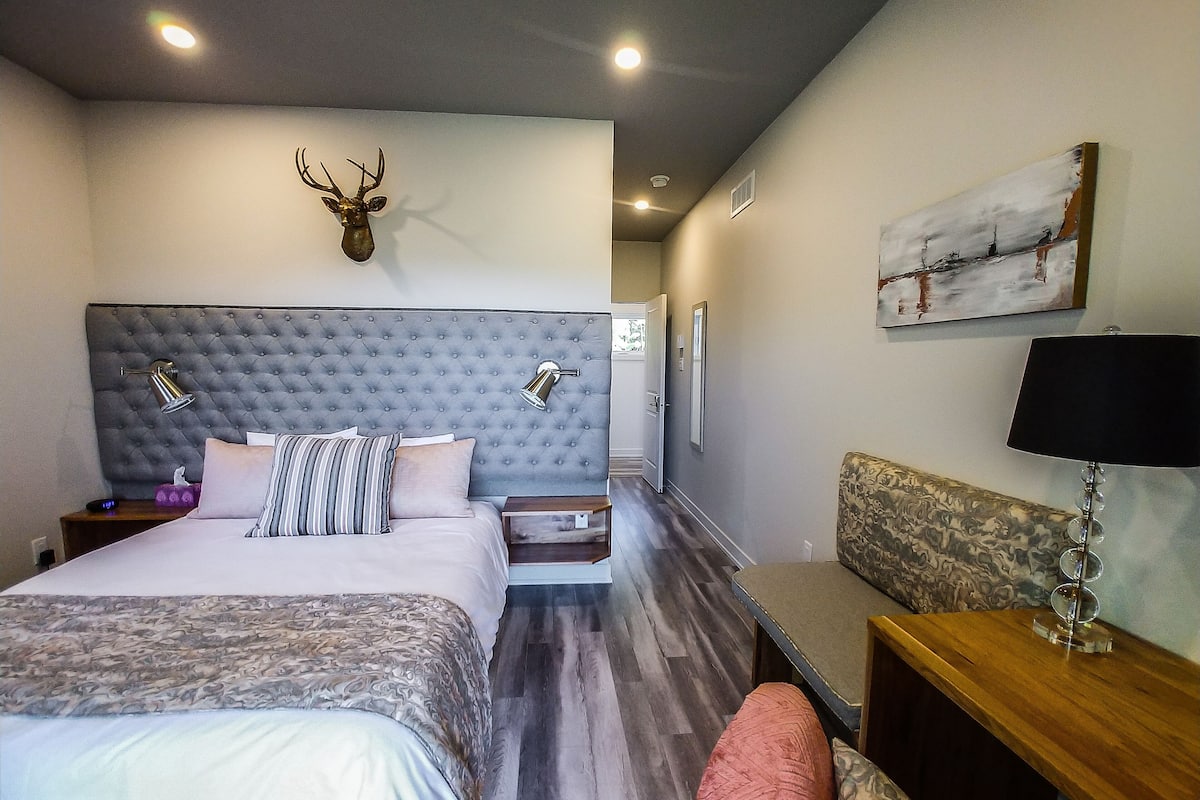 A hotel room with a tufted headboard, a mounted deer head, a bed with patterned bedding, a bedside table with a lamp, a sofa, and a painting on the wall.