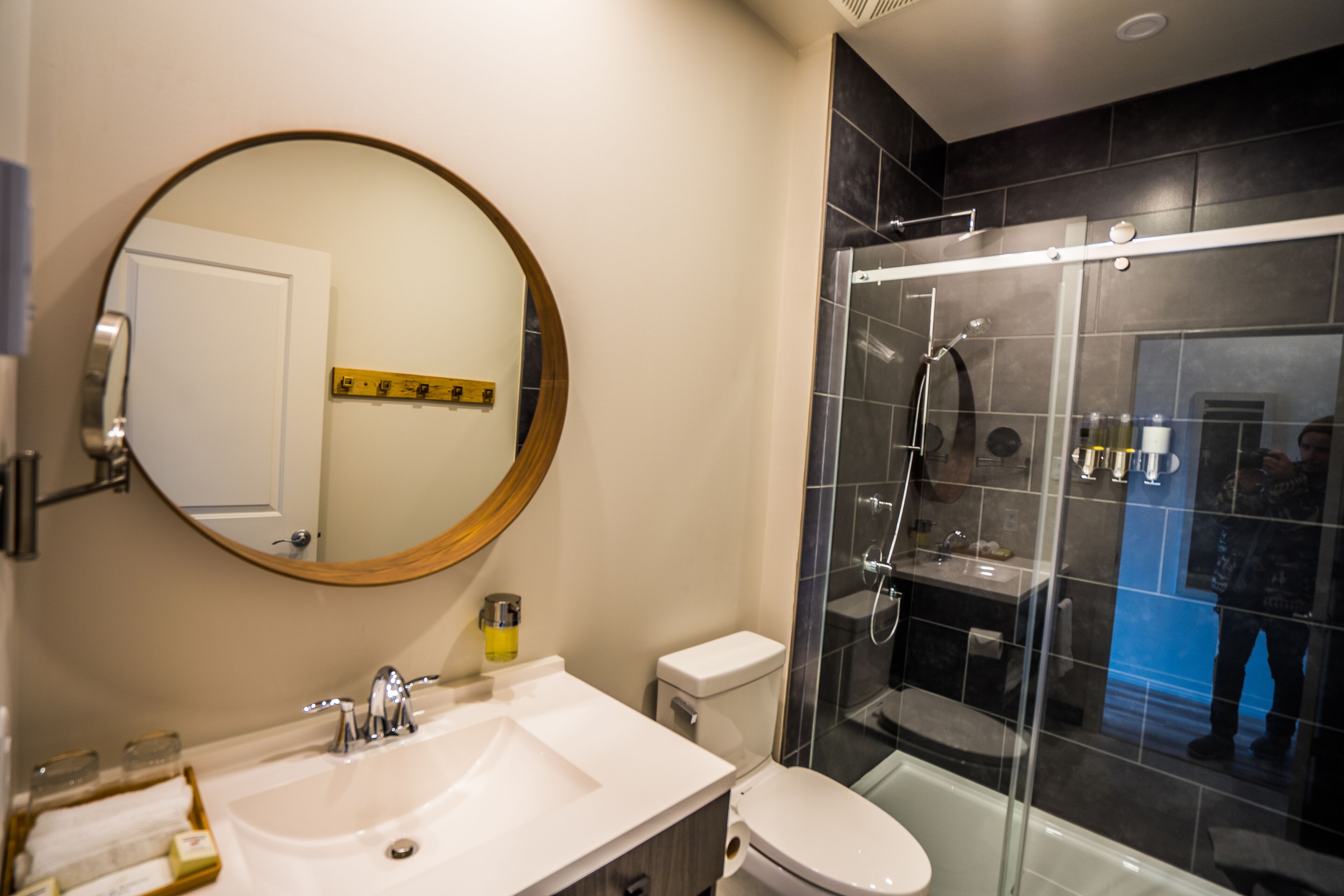 Un baño moderno con un espejo redondo, un lavamanos blanco, un inodoro y una zona de ducha con mampara de vidrio.