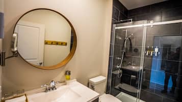 Un baño moderno con un espejo redondo, un lavamanos blanco, un inodoro y una zona de ducha con mampara de vidrio.