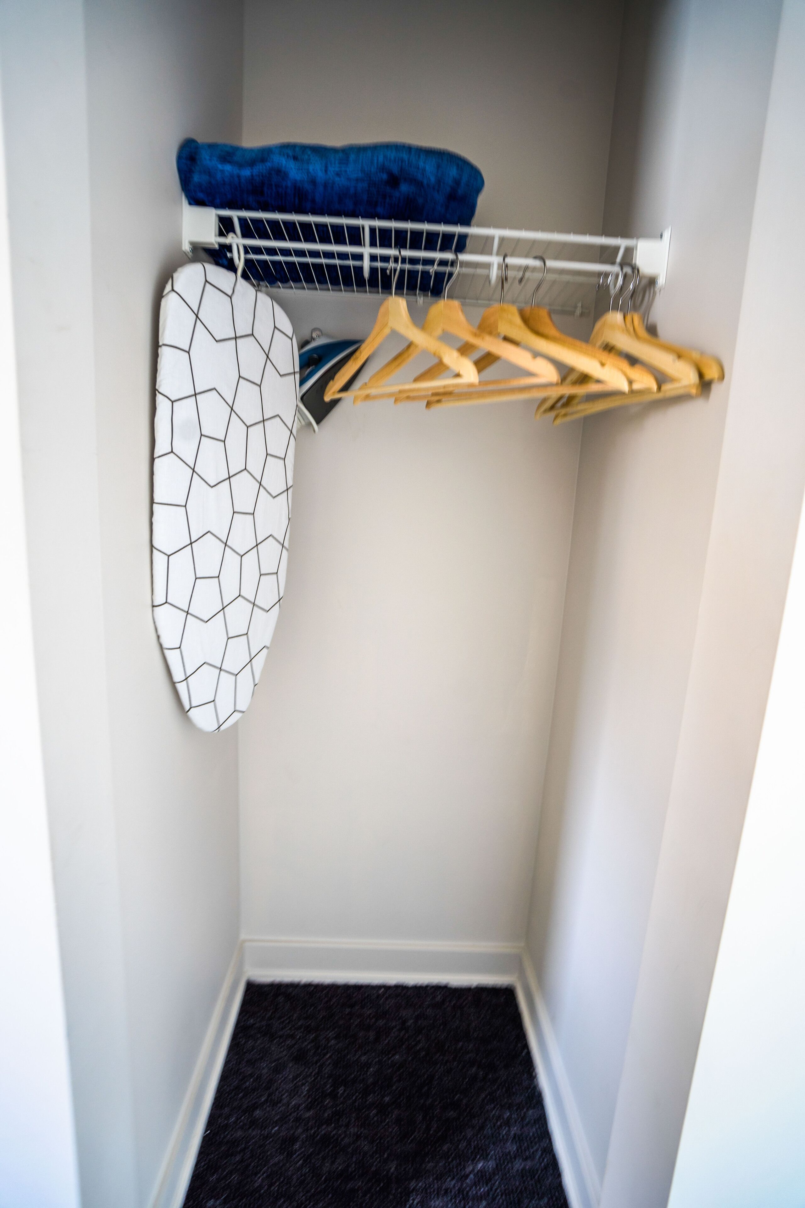 Un espace de rangement compact doté d’une tringle pour suspendre les vêtements, d’une planche à repasser blanche à motifs géométriques et d’un sol recouvert d’une moquette sombre.