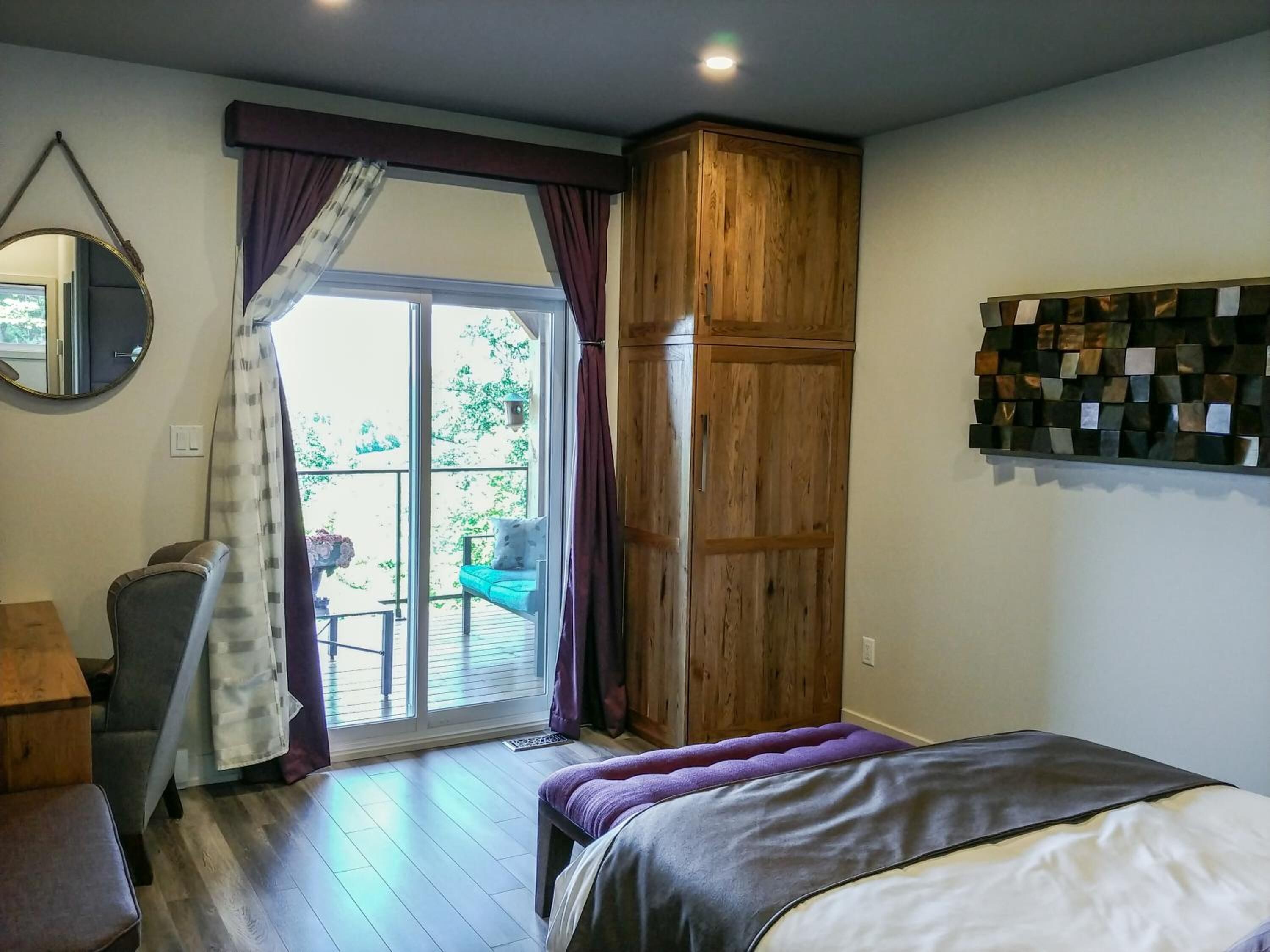 Une chambre à coucher avec un lit, une armoire en bois, une chaise, un bureau et une grande fenêtre avec des rideaux.