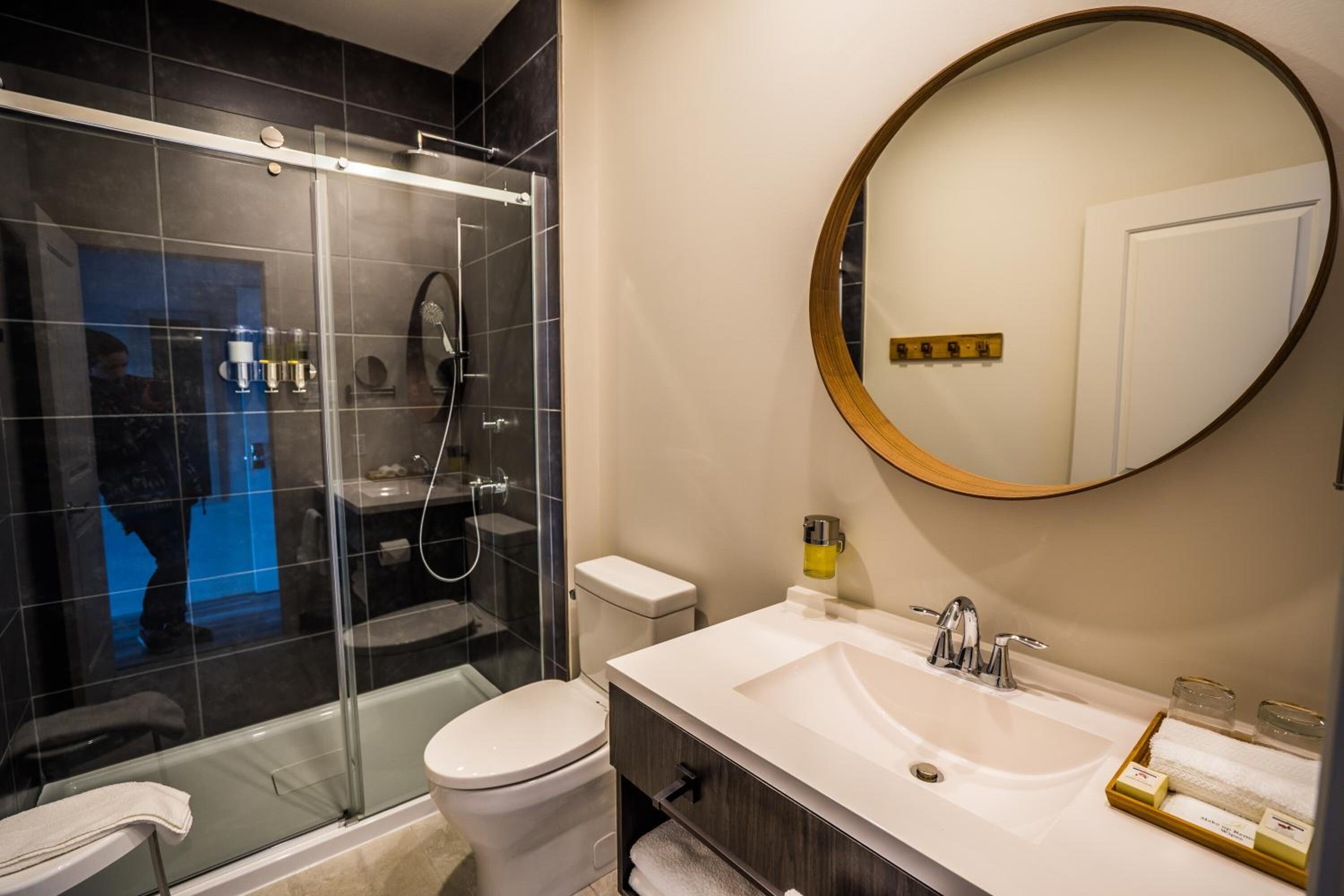 A modern bathroom with a glass-enclosed shower, a white toilet, a large round mirror, and a white sink with a wooden cabinet.
