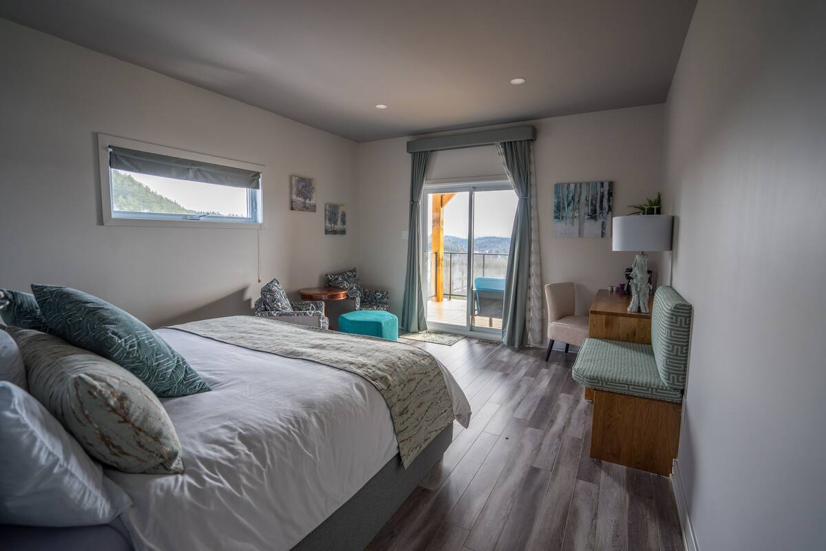 A bedroom with a bed, a chair, a table, a lamp, and a view of the outdoors.