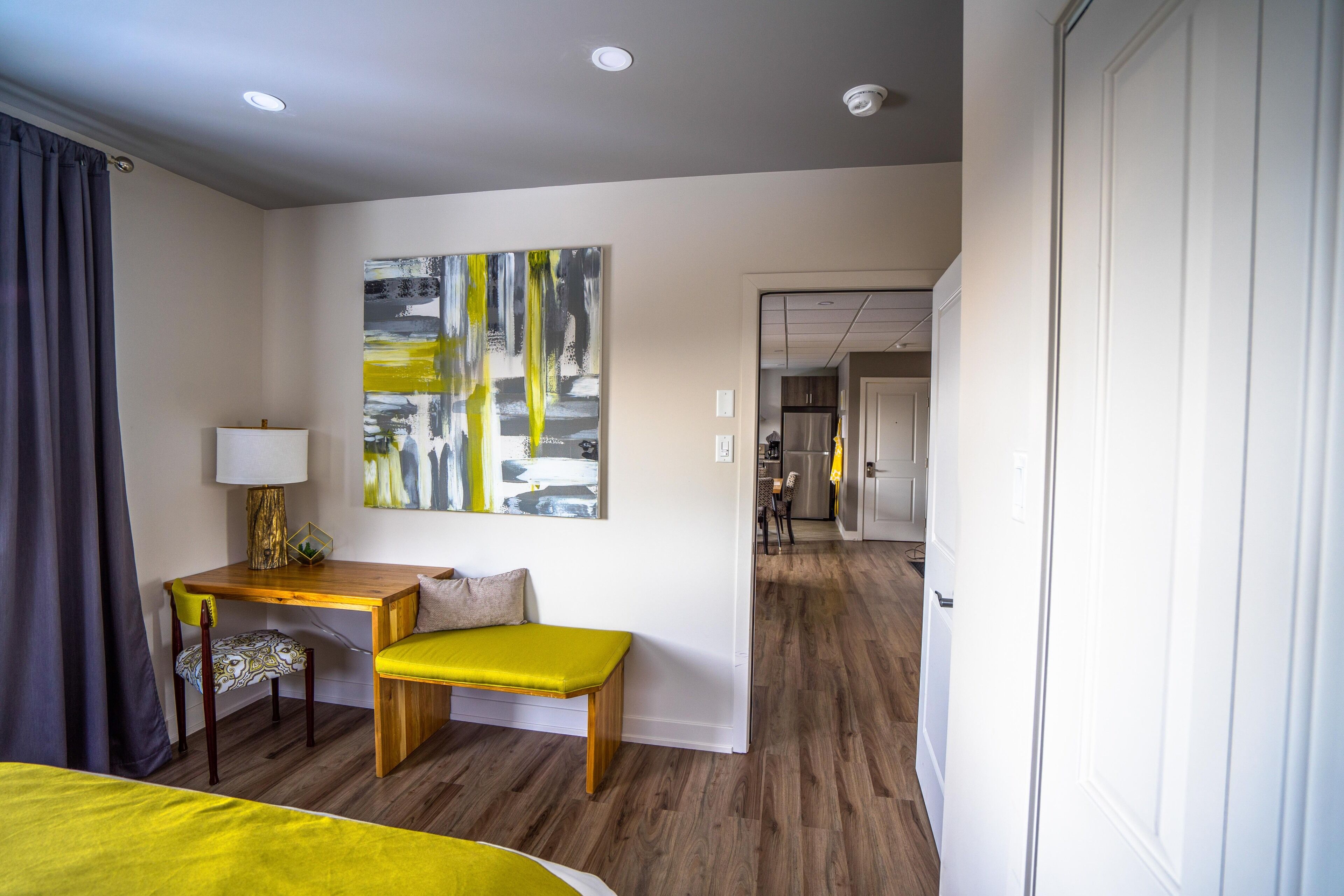 A hotel room with a bed, a desk with a lamp, a bench, a painting on the wall, and a view into the kitchen area.