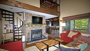 A spacious living room with a fireplace, a staircase, a kitchen in the background, and a large window providing a view of the outdoors.