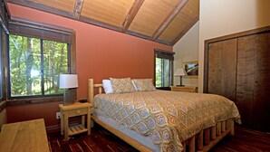 A bedroom with a wooden bed, a nightstand with a lamp, a window with a view of trees, and a wooden wardrobe.