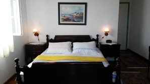 A neatly made bed with white and yellow bedding, two bedside tables with lamps, a framed painting of boats on the wall, and a view of a door and hallway. - Lake House Oasis, Dodoši, Skadar lake (Dodoši)