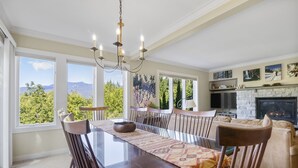 A dining area with a large table, chairs, and a chandelier. There is a fireplace with a stone surround and a television mounted above it. Large windows provide a view of the outdoors. - Topnotch Overlook Resort Home with Mansfield views (Stowe)