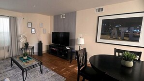 A living room with a flat-screen TV, a dining table with chairs, a coffee table, and a large window with curtains.