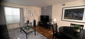 A living room with a flat-screen TV, a dining table with chairs, a coffee table, and a large window with curtains. - Walk to Aquarium & Stadiums · Free Parking (Atlanta)