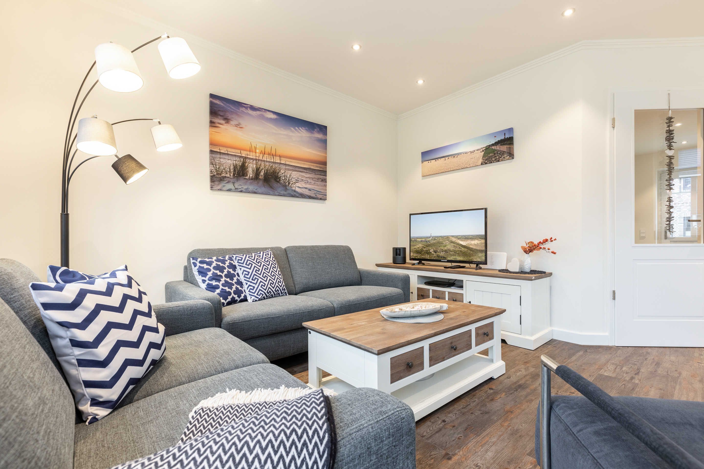 Ein modernes Wohnzimmer mit grauem Sofa, einem Couchtisch und einem Fernseher. An den Wänden hängen Bilder, und eine Stehlampe steht im Raum.