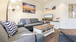 Una sala de estar moderna con un sofá gris, una mesa de centro y un televisor. Hay cuadros colgados en la pared y una lámpara de pie.