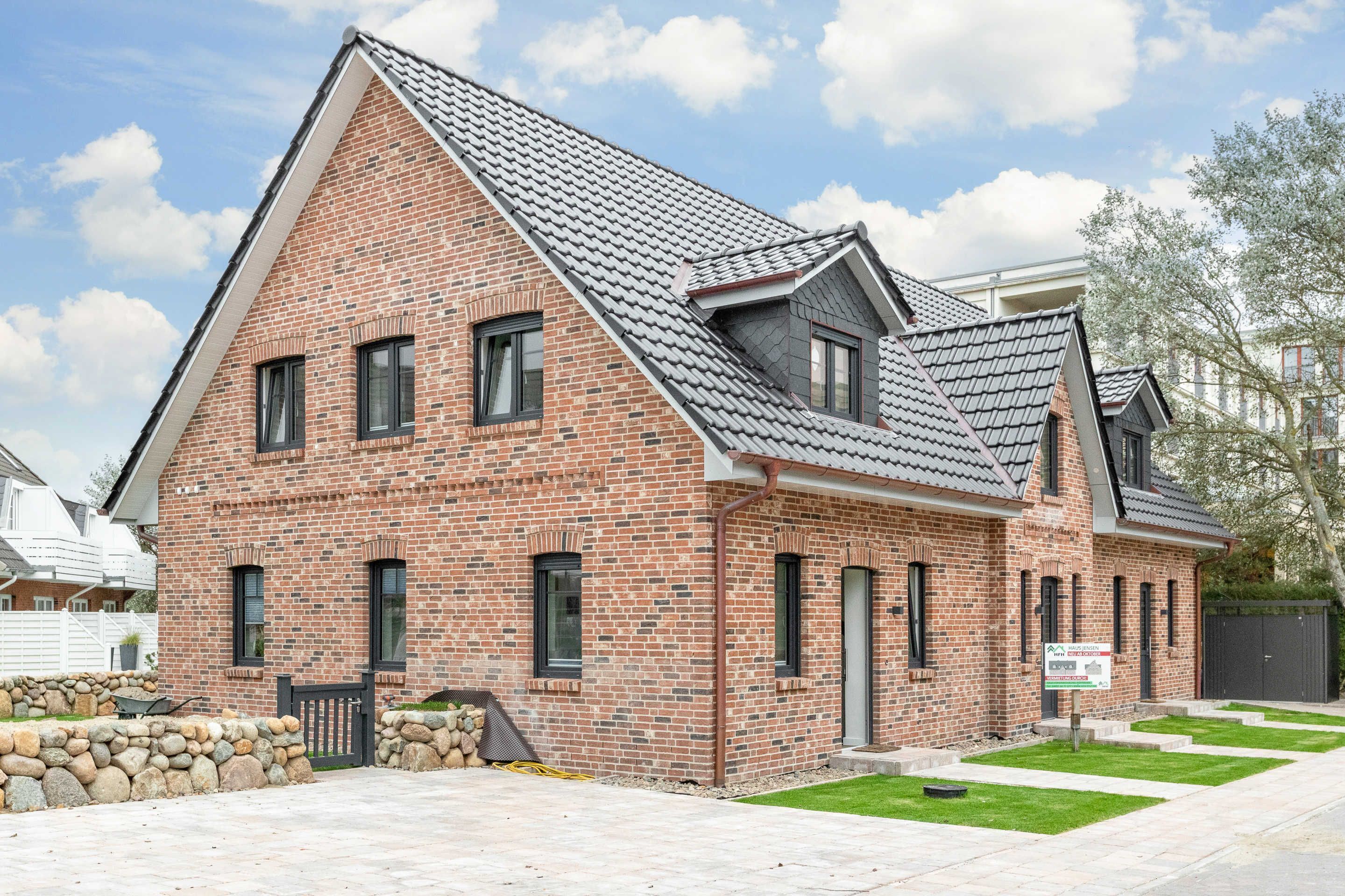 A two-story brick house with a sloped roof, a stone wall, and a small front yard.