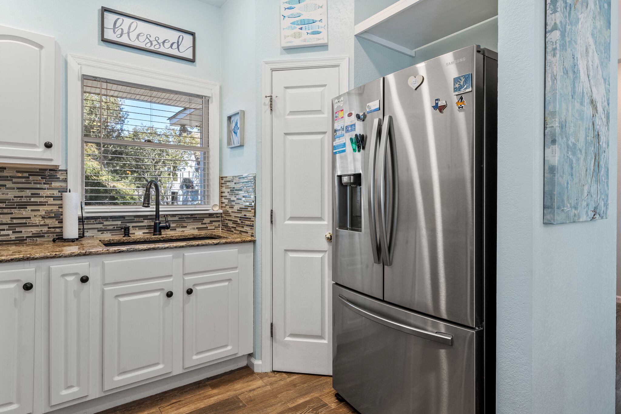 A kitchen with white cabinets, a stainless steel refrigerator, a window with a view, and a 'blessed' sign on the wall.