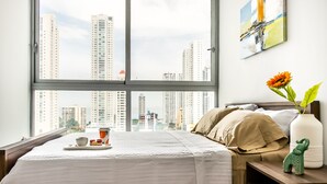 A hotel room with a large window, a bed with a white bedspread, a nightstand with a vase of flowers, and a painting on the wall. - Panoramic Apartment Ocean View (Panama City)