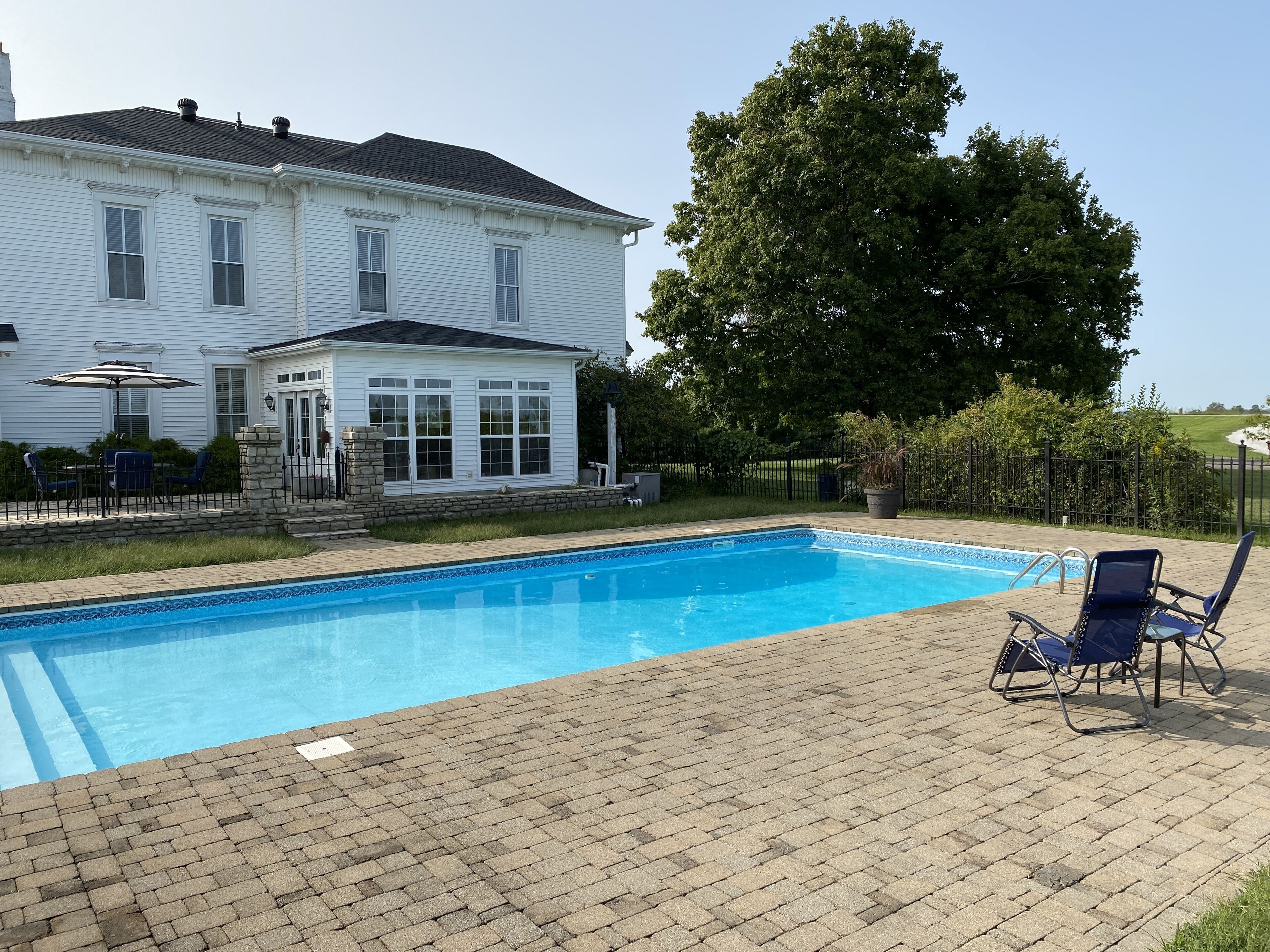 A rectangular swimming pool with a brick deck, surrounded by a white house with a covered patio and outdoor furniture.