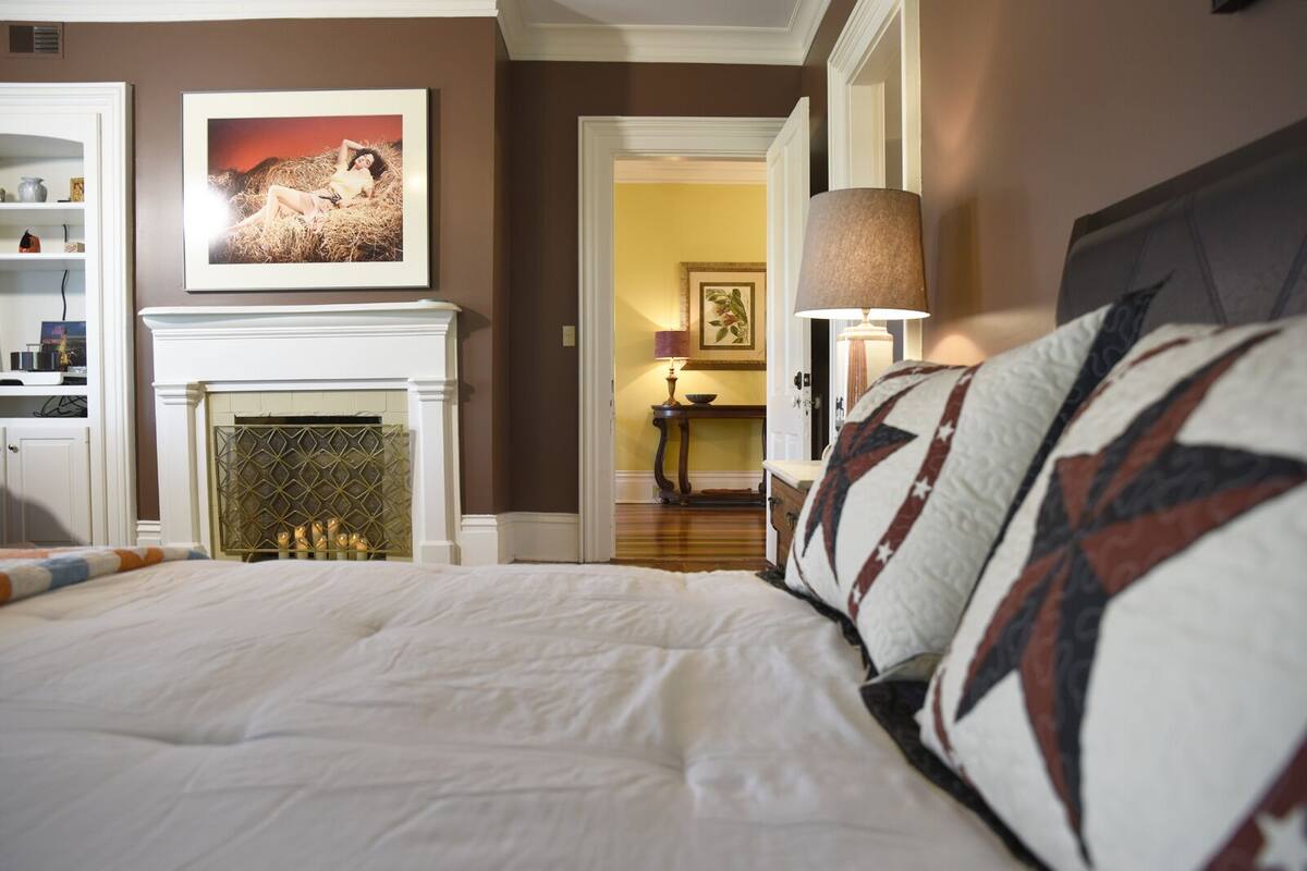 A hotel room with a large bed, a fireplace, a framed picture on the wall, and a lamp.