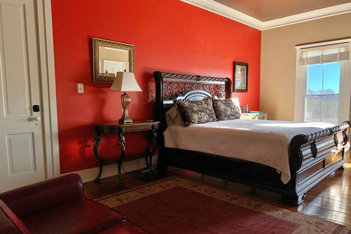 A bedroom with a red wall, a bed with a dark headboard, a nightstand, a lamp, a mirror, and a red leather chair.