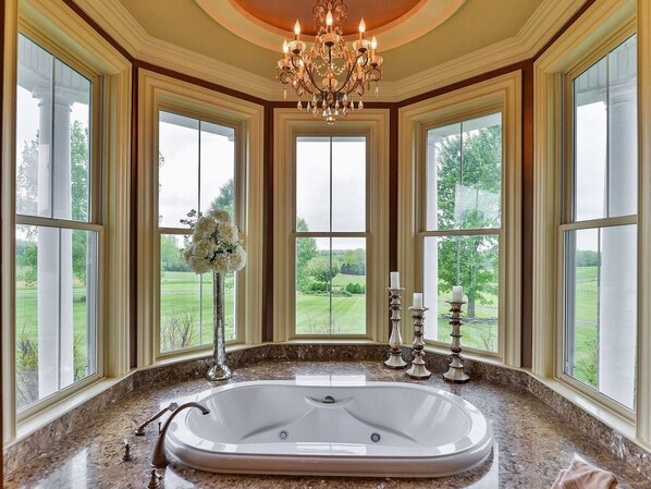 A spacious bathroom with a large bathtub, multiple windows offering a view of a green landscape, and a classic chandelier.