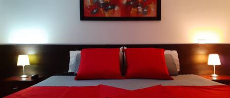A hotel room with a bed, red and white pillows, two bedside lamps, and a framed abstract painting on the wall.