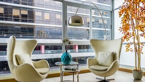Un intérieur moderne avec deux fauteuils à oreilles, une petite table ronde, un lampadaire et une grande fenêtre donnant sur des immeubles.