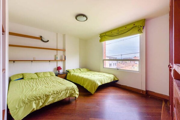 A small bedroom with two single beds, a wooden shelf, a small table, and a window with a view of buildings. - La Macarena 2001 (Bogotá)