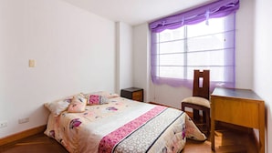 A bedroom with a bed, a chair, a small table, and a window with a purple curtain. - La Macarena 2001 (Bogotá)