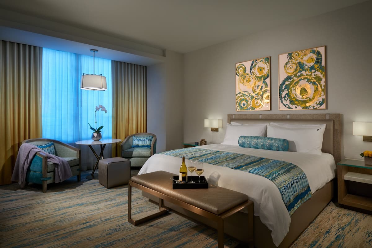 Habitación de hotel con una cama grande, dos mesitas de noche con lámparas, un banco, una pequeña mesa con una planta y una silla.
