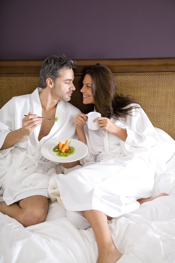 A couple in white bathrobes sitting on a bed with a plate of food and a cup.