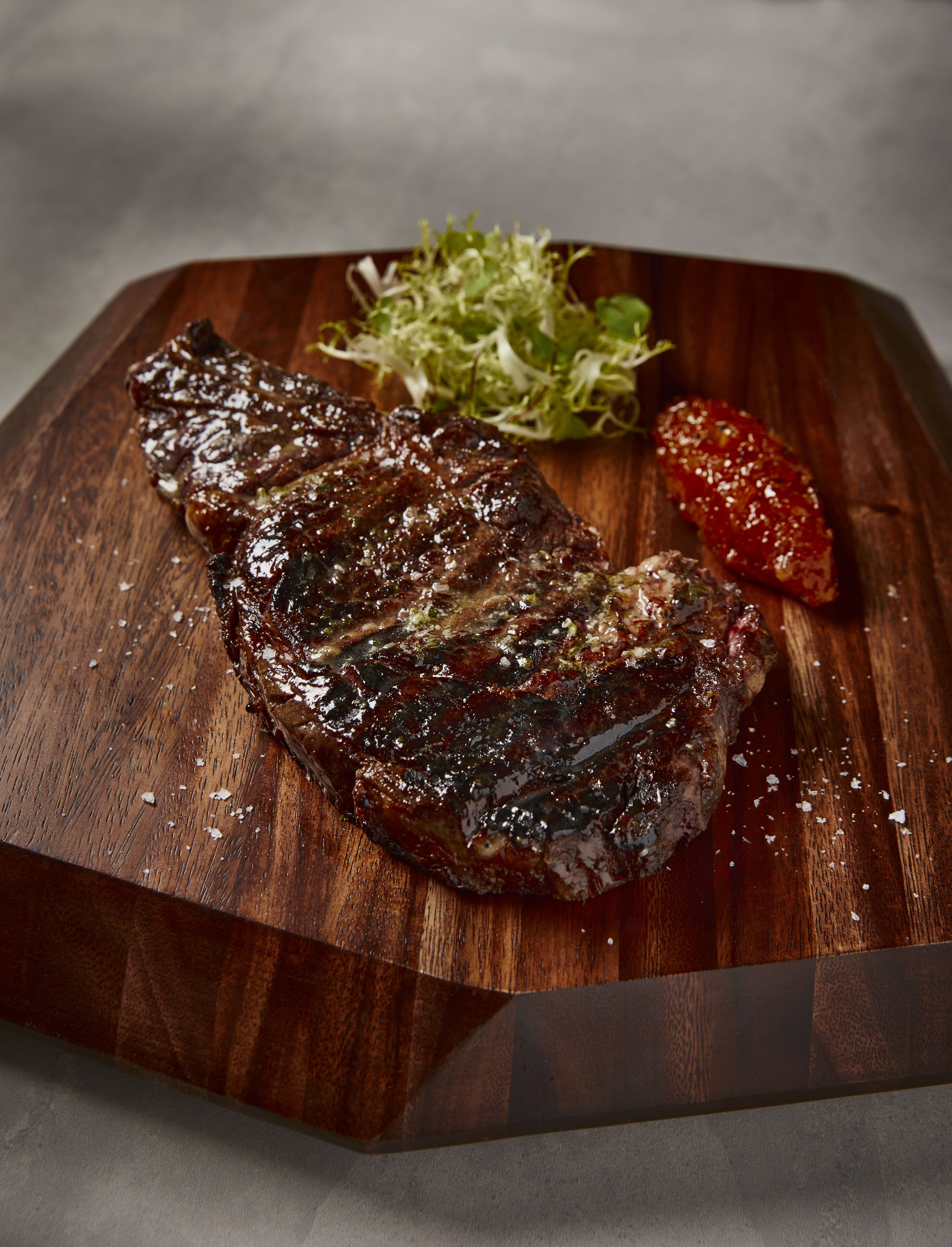 A steak on a wooden cutting board with a side of greens and a slice of tomato.
