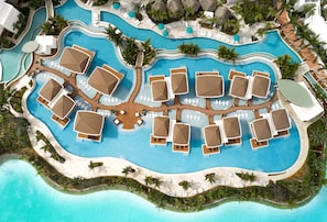 An aerial view of a resort with a large central pool, multiple sun loungers, and surrounding palm trees.