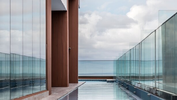 Una piscina infinita moderna con barandillas de vidrio y estructura de columnas de madera, con vistas a un paisaje distante.