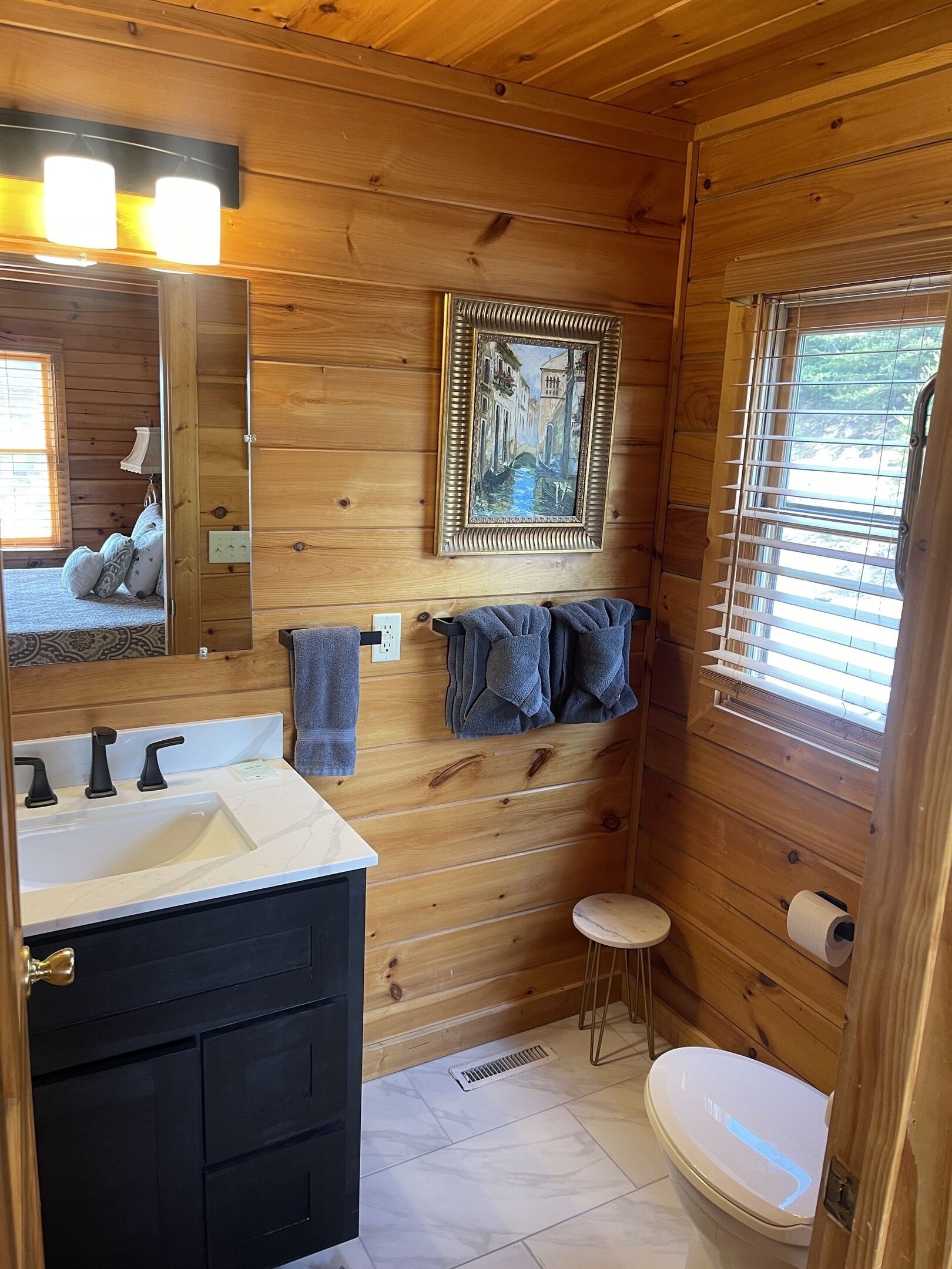 A wooden cabin bathroom with a sink, mirror, towel rack, toilet, and a window with blinds.