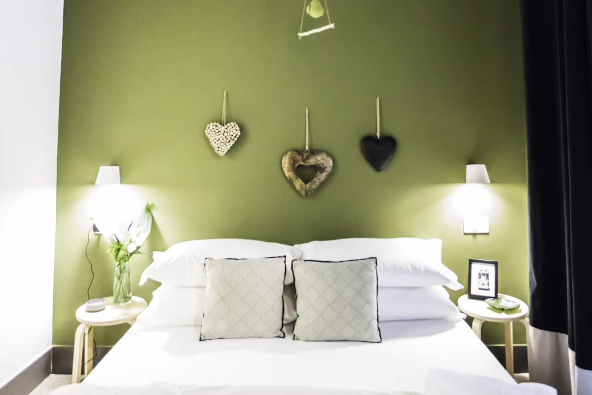 A neatly made bed with white linens and two pillows, a bedside table with a lamp, and heart-shaped wall decorations.