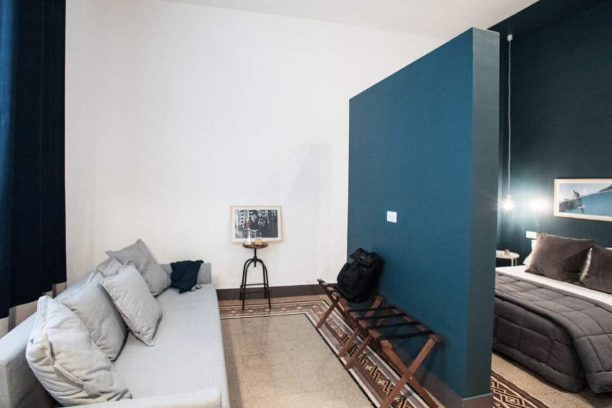 A hotel room with a bed, a sofa, a small table with a picture, and a chair.