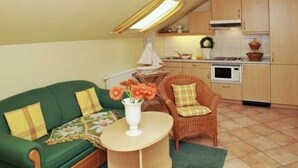 A cozy living area with a sofa, a wicker chair, a small table with flowers, and a kitchen in the background.