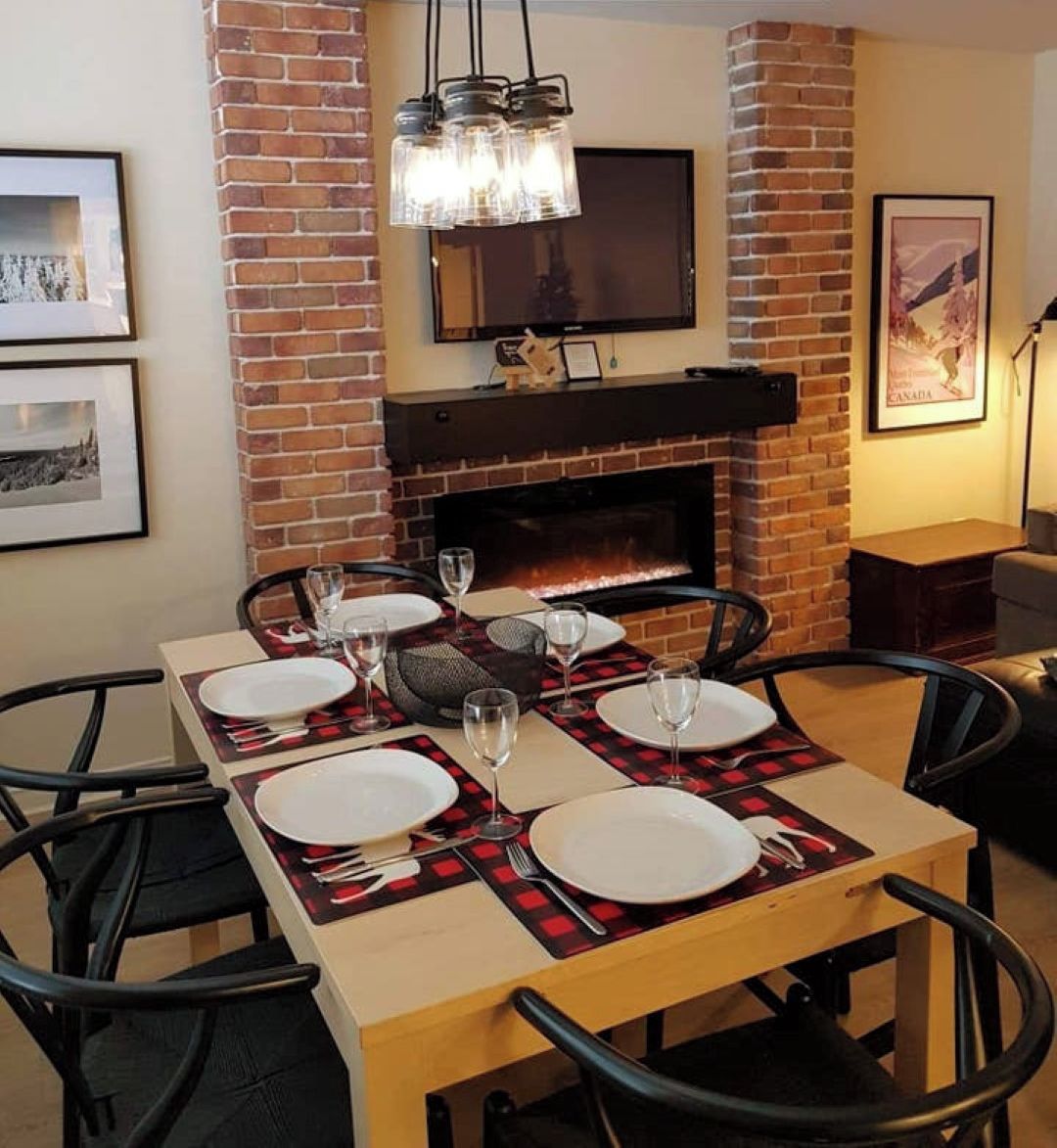 A dining area with a brick fireplace, a television mounted above it, and a table set for four with wine glasses and plates.