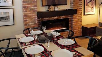 A dining area with a brick fireplace, a television mounted above it, and a table set for four with wine glasses and plates.
